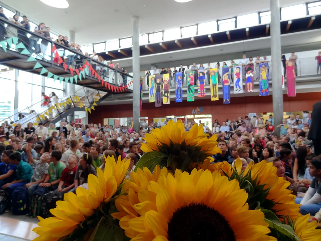 Sonnenblumenstrauß im Vordergrund, dahinter die gefüllte Aula.